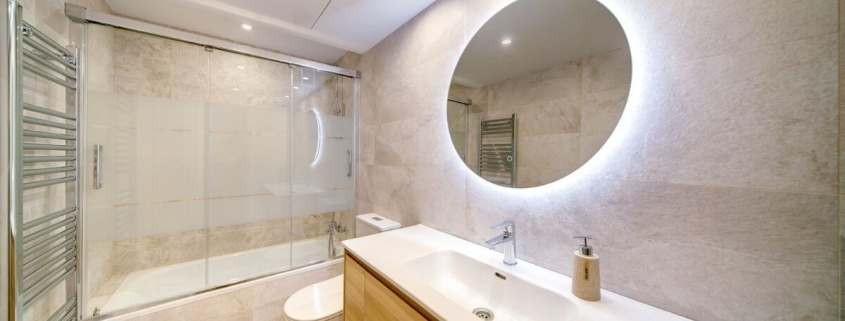 Baño moderno reformado en Zaragoza con diseño elegante, espejo circular retroiluminado, lavabo de madera y bañera con mampara de cristal